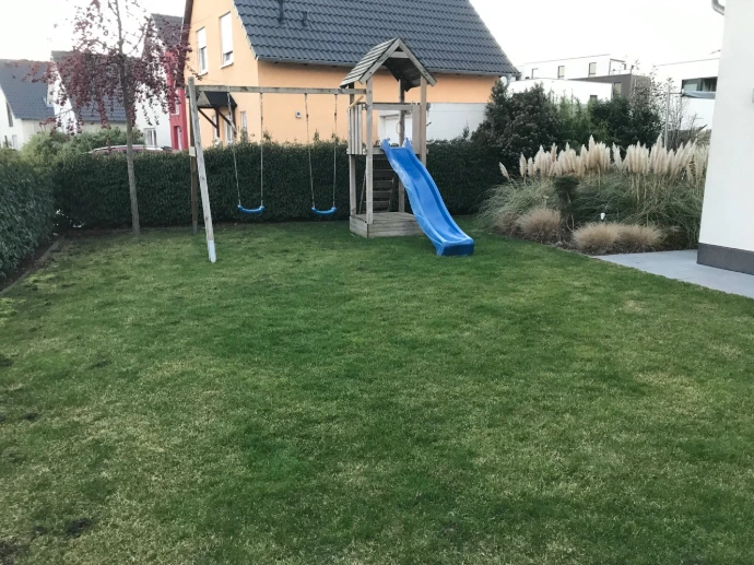 Hausgarten mit frisch angelegtem Rasen, Schaukel und Rutsche vor Gräsern und Hecke